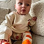 baby, child, pajamas, orange_socks, pumpkin_pattern, couch, pillows, beige, soft_texture, indoor, cute, curious, sitting, clothing, face, person, young_child, cozy, home, portrait