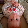baby, infant, onesie, clothing, red_letters, heart_pattern, bed, blanket, lying_down, hands_folded, looking_up, face, head, skin, person, cute, cozy, indoors, sleepwear, child