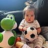 baby, child, toy, dinosaur_toy, soccer_ball, blanket, couch, indoor, cute, portrait, infant, soft_lighting, playtime, casual_clothing, person, seated, expression, hair_tie, red_bracelet, floor