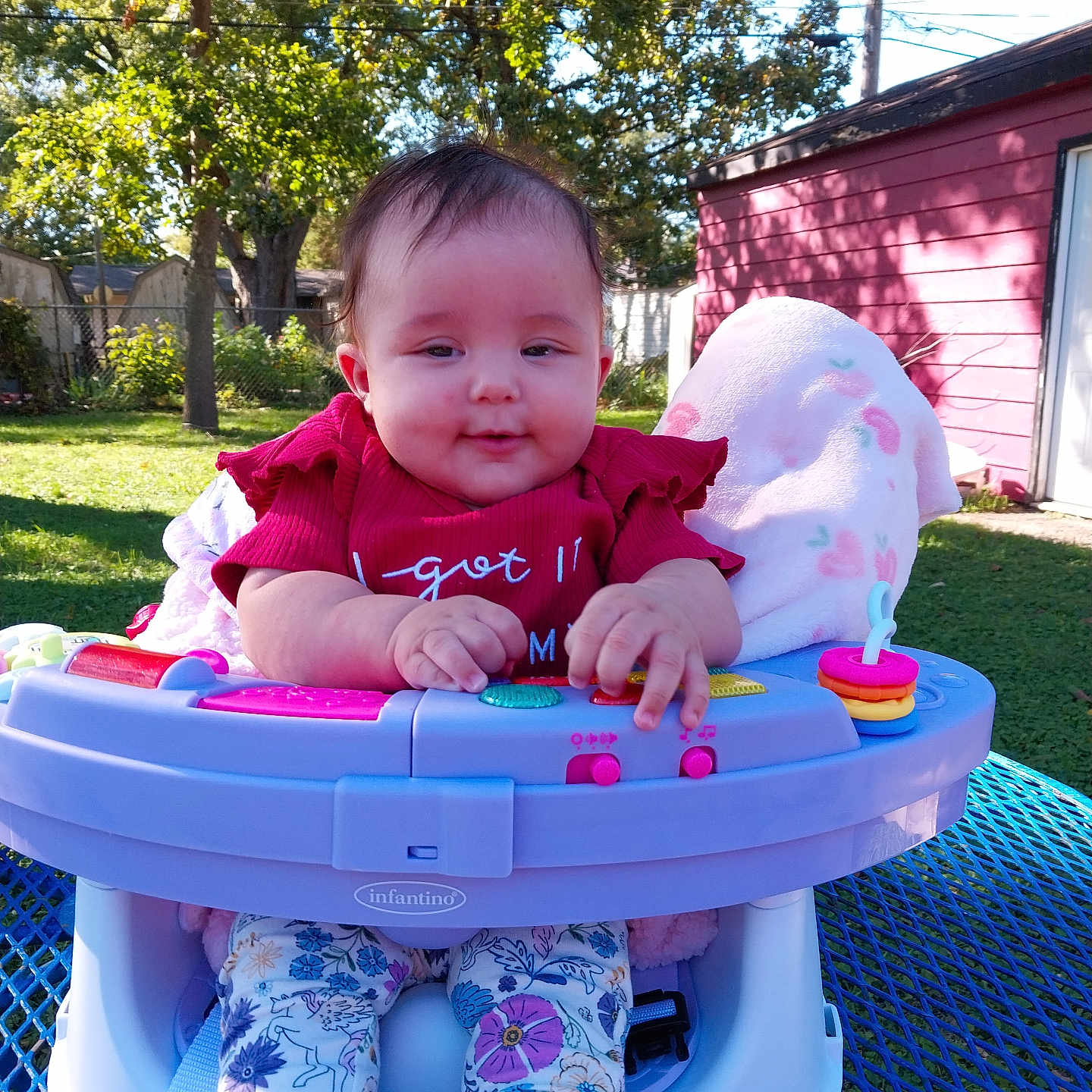 Sophia is registered to the contest to win money with this photo: baby, child, infant, outdoor, backyard, activity_center, jumper, picnic_table, smiling, face, portrait, red_shirt, floral_pants, toy, colorful, trees, grass, shed, sunlight, family_photo