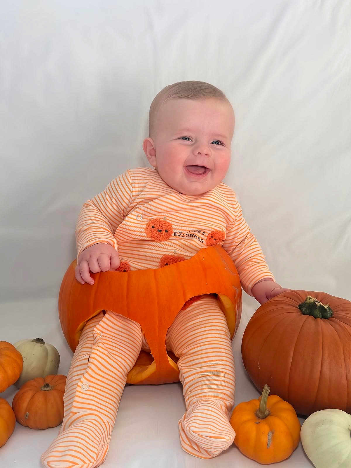 Frankie is registered to the contest to win money with this photo: baby, pumpkin, orange_striped_pajamas, smiling, halloween, holiday, cute, child, festive, autumn, costume, celebration, sitting, happy, face, person, small_pumpkins, white_background, indoors, fun