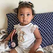 Reigné joined the competition — help win amazing prizes! toddler, child, baby, curly_hair, pigtails, white_onesie, bunny_embroidered, blanket, pillow, patterned_pillow, indoor, sitting, cute, portrait, face, hand, toy, pacifier_clip, soft_lighting, wall