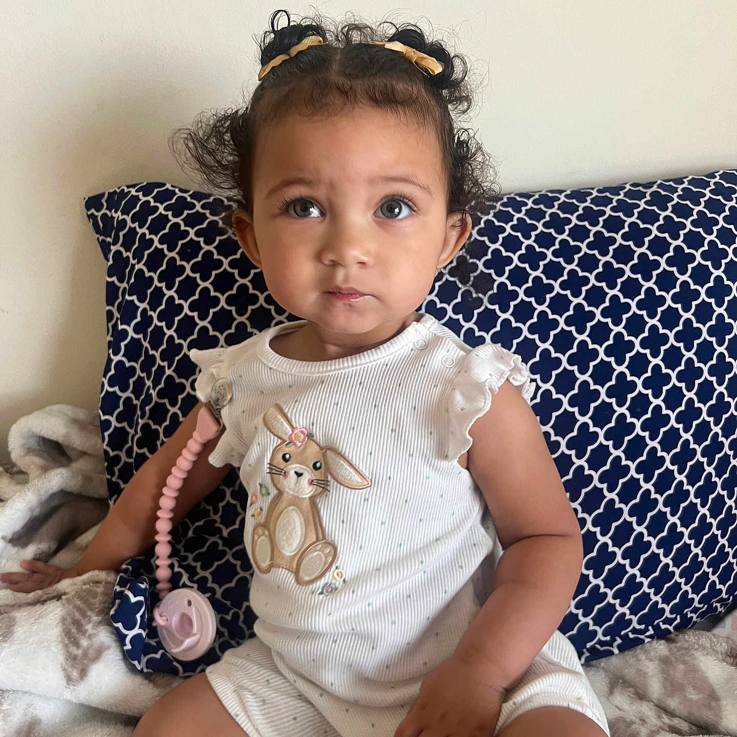 Reigné joined the competition — help win amazing prizes! baby, blanket, bunny_embroidered, child, curly_hair, cute, face, hand, indoor, pacifier_clip, patterned_pillow, pigtails, pillow, portrait, sitting, soft_lighting, toddler, toy, wall, white_onesie