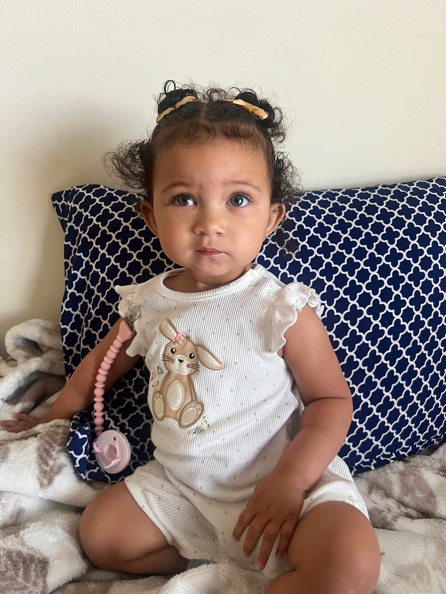 Reigné joined the competition — help win amazing prizes! toddler, child, baby, curly_hair, pigtails, white_onesie, bunny_embroidered, blanket, pillow, patterned_pillow, indoor, sitting, cute, portrait, face, hand, toy, pacifier_clip, soft_lighting, wall