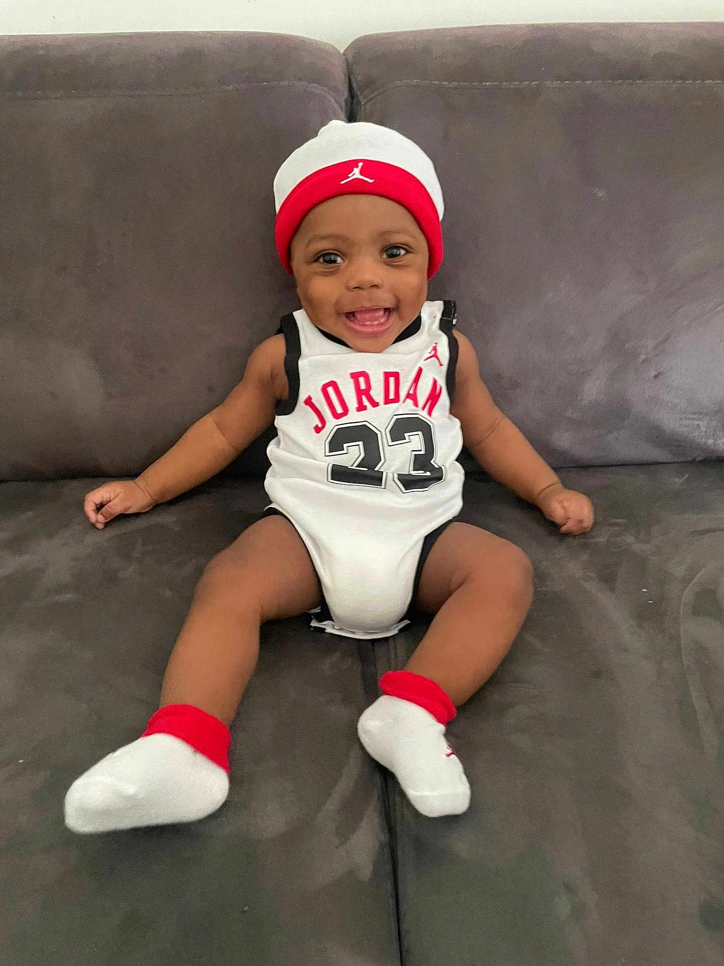 Kyron-Mace participe au concours pour gagner de l'argent avec cette photo : baby, infant, smiling, sitting, sofa, couch, white_onesie, jersey, number_23, jordan, hat, beanie, socks, happy, portrait, indoors, living_room, child, hands, feet