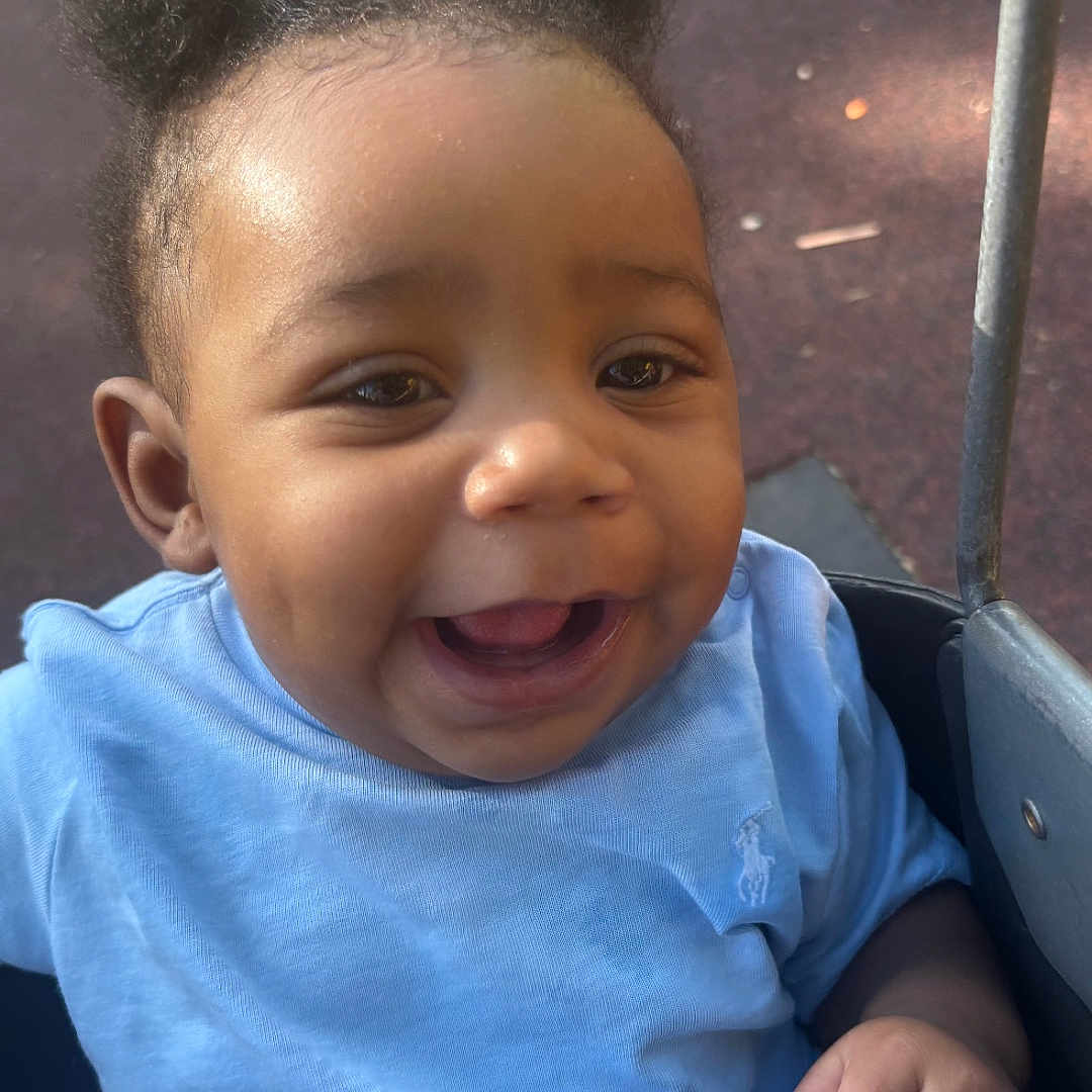 Zumir joined the competition — help win amazing prizes! baby, blue_shirt, brown_skin, child, cute, daylight, face, fist, hair_buns, hand, happy, infant, looking_at_camera, open_mouth, outdoor, playground, portrait, seated, smiling, stroller