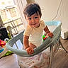 Maverick is registered to the contest to win money with this photo: toddler, child, baby, playpen, indoor, wood_floor, window, light, smile, curly_hair, white_clothing, bracelet, blanket, toy, home, furniture, person, cute, happy, babygear