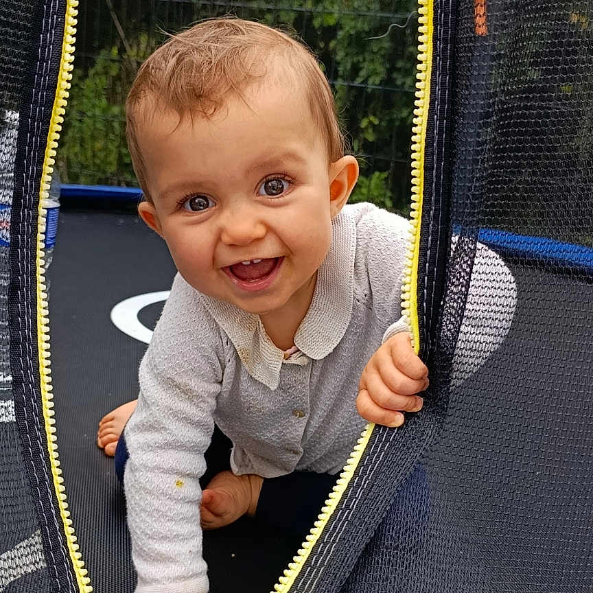 Zeylïnah a rejoint le concours — aidez-le/la à gagner de superbes lots ! baby, barefoot, child, clothing, crawling, curious, fence, greenery, happy, infant, long_sleeve, mesh, nature, outdoor, playful, smiling, trampoline, yellow_zipper, young_child, zipper