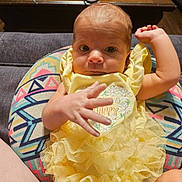 Payton is registered to the contest to win money with this photo: baby, infant, child, yellow_dress, ruffles, cushion, patterned, hand, face, eyes, indoor, furniture, wooden_table, curious, person, young, cute, sitting, colorful, soft