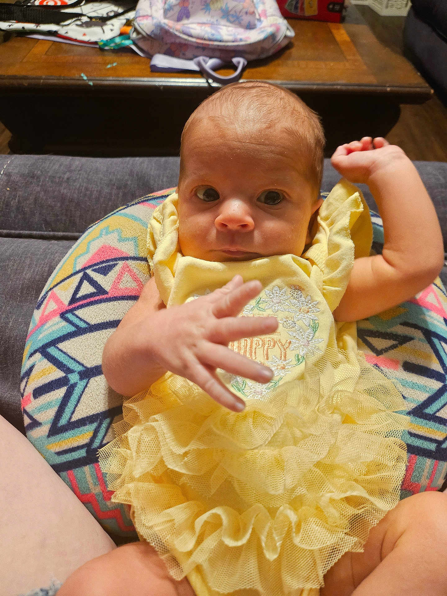 Payton is registered to the contest to win money with this photo: baby, infant, child, yellow_dress, ruffles, cushion, patterned, hand, face, eyes, indoor, furniture, wooden_table, curious, person, young, cute, sitting, colorful, soft