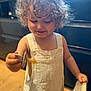 toddler, child, curly_hair, overalls, indoor, spoon, food, cloth, smile, happy, person, baby, light, wooden_floor, cabinet, play, cute, small, standing, apparel