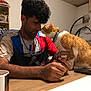 beaded_necklace, cards, casual_clothing, cat, cup, friendly_interaction, game, indoor, lamp, man, orange_cat, pet, playing_cards, shelf, sitting, smiling, table, uno, white_cat, wooden_table