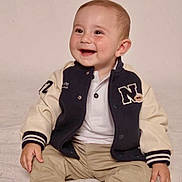 Christopher joined the competition — help win amazing prizes! baby, child, smile, sitting, jacket, pants, footwear, happy, portrait, indoor, cute, young, infant, clothing, person, face, head, warm, soft, blanket
