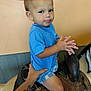 toddler, child, blue_shirt, shorts, saddle, hand, tattoo, indoor, curious_expression, pout, person, leather, horse_saddle, seated, furniture, skin, short_hair, background, colorful_wall, holding