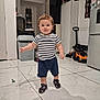 toddler, child, indoor, striped_shirt, navy_shorts, shoes, tile_floor, pacifier, toy_car, kitchen, refrigerator, cabinet, trash_can, shadow, person, young_child, cute, standing, home, furniture