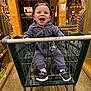 baby, child, shopping_cart, cap, smiling, striped_clothing, footwear, indoor, store, wooden_pillars, shelves, retail, happy, person, infant, flooring, toy, pacifier, casual, shopping