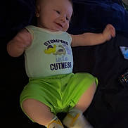 Cayson is registered to the contest to win money with this photo: baby, infant, child, smiling, clothing, onesie, socks, green_shorts, bed, blanket, remote_control, happy, cute, lying_down, indoor, person, face, young_child, movement, soft_lighting