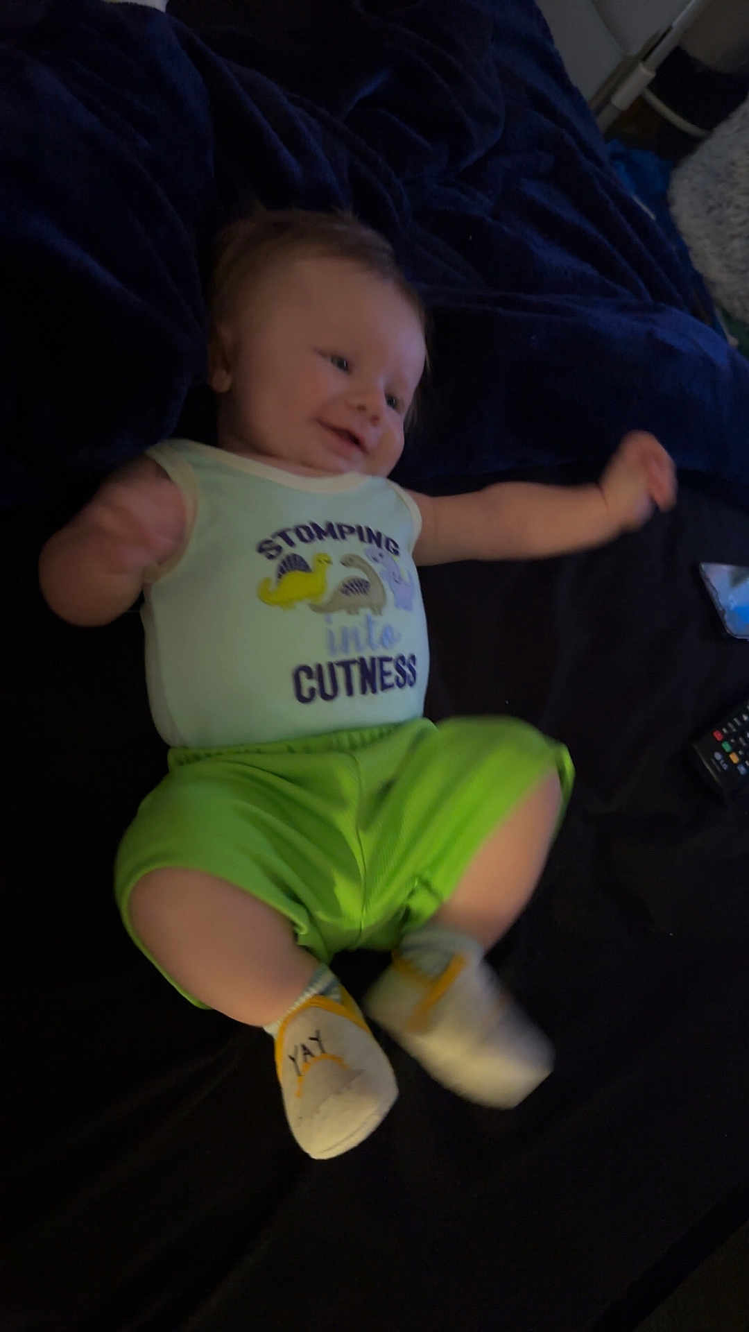 Cayson is registered to the contest to win money with this photo: baby, infant, child, smiling, clothing, onesie, socks, green_shorts, bed, blanket, remote_control, happy, cute, lying_down, indoor, person, face, young_child, movement, soft_lighting