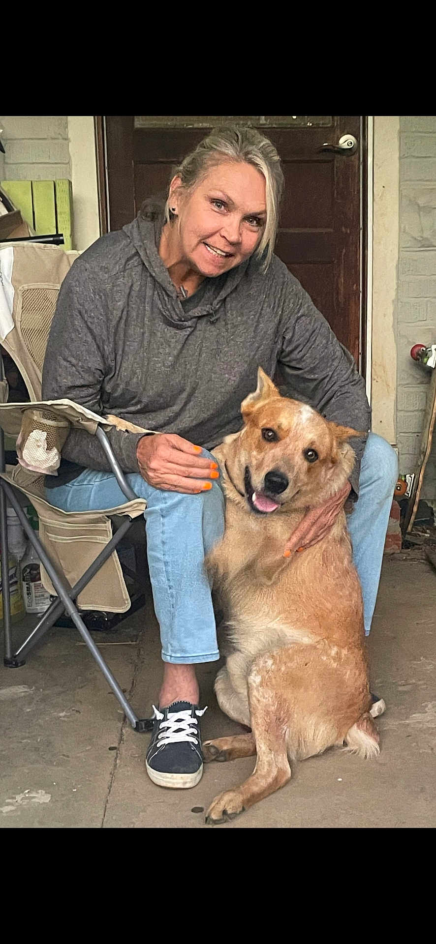 Hank is registered to the contest to win money with this photo: woman, dog, pet, smiling, sitting, folding_chair, porch, casual_clothing, gray_hoodie, light_blue_jeans, sneakers, orange_nail_polish, happy_dog, tongue_out, wooden_door, brick_wall, concrete_floor, indoor, affection, human_animal_interaction