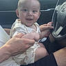 baby, child, car, steering_wheel, smile, happy, person, hands, sitting, clothing, shorts, seat, window, daylight, vehicle, interior, nature, trees, white_car, portrait