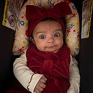 Carolina is registered to the contest to win money with this photo: baby, infant, child, car_seat, headband, bow, red_clothing, white_sleeves, floral_pattern, face, wide_eyes, hand, cute, portrait, indoor, warning_label, fabric, seat, expression, skin