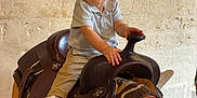 Keylan participe au concours pour gagner de l'argent avec cette photo : toddler, child, saddle, indoor, stone_wall, western_saddle, light_blue_shirt, khaki_pants, white_sneakers, sitting, curious, young_child, leather, horse_tack, furniture, floor, hand, portrait, person, baby