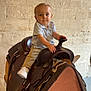 toddler, child, saddle, indoor, stone_wall, western_saddle, light_blue_shirt, khaki_pants, white_sneakers, sitting, curious, young_child, leather, horse_tack, furniture, floor, hand, portrait, person, baby