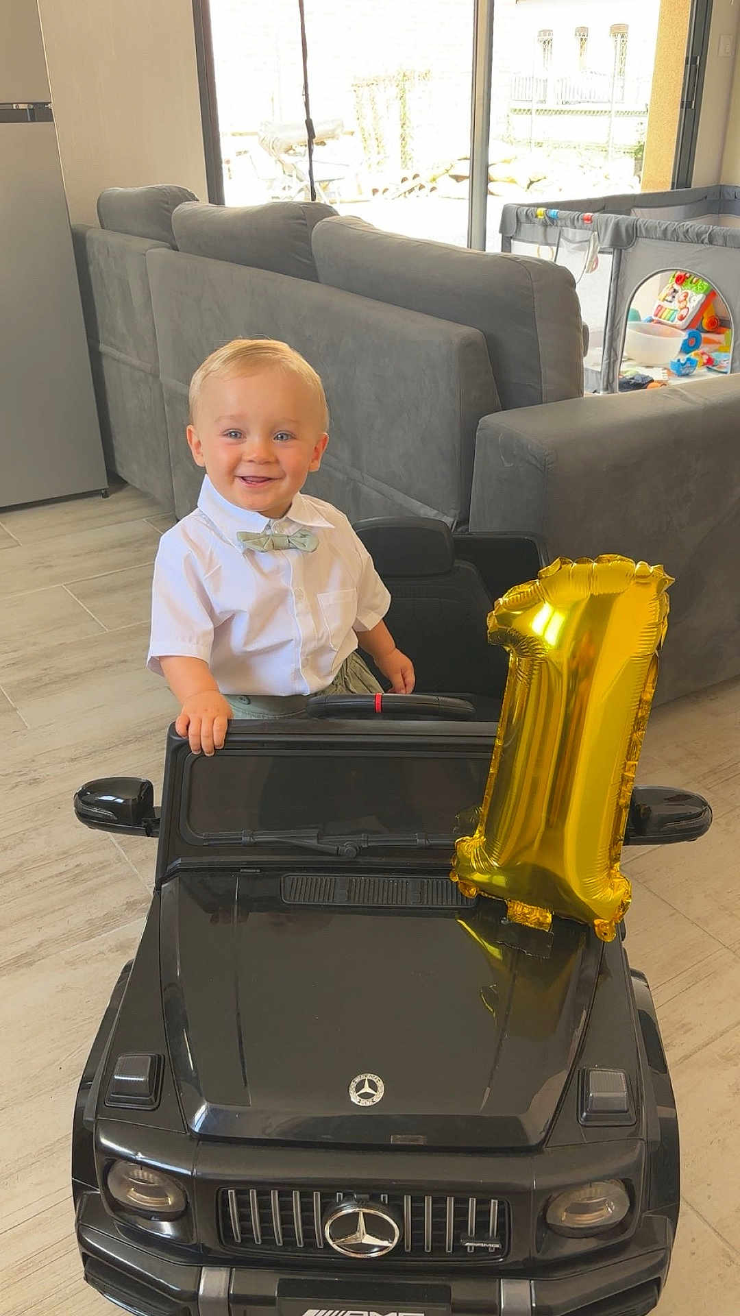 Keylan a rejoint le concours — aidez-le/la à gagner de superbes lots ! toddler, child, toy_car, birthday_balloon, number_one_balloon, indoor, living_room, sofa, playpen, toy, smiling, bow_tie, shirt, floor, window, daylight, happy, person, celebration, blond_hair