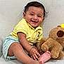 baby, child, smiling, yellow_shirt, striped_shorts, teddy_bear, plush_toy, barefoot, sitting, happy, cute, indoor, portrait, infant, playful, hand_touching_foot, soft_texture, light_background, young, person