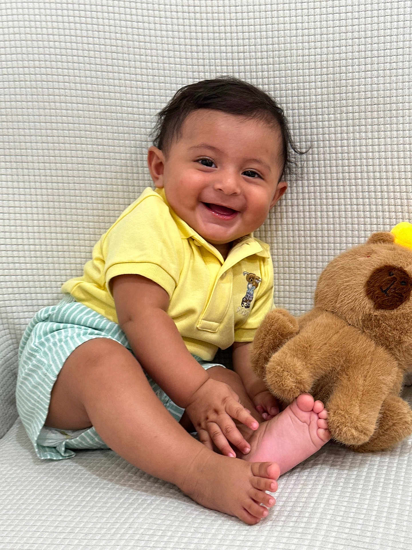 Gabriel joined the competition — help win amazing prizes! baby, child, smiling, yellow_shirt, striped_shorts, teddy_bear, plush_toy, barefoot, sitting, happy, cute, indoor, portrait, infant, playful, hand_touching_foot, soft_texture, light_background, young, person
