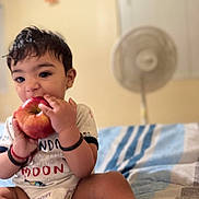 Faivish is registered to the contest to win money with this photo: toddler, child, apple, bite, bed, blanket, indoor, person, cute, happy, clothing, bracelet, fan, furniture, home, portrait, sitting, young_child, face, playful