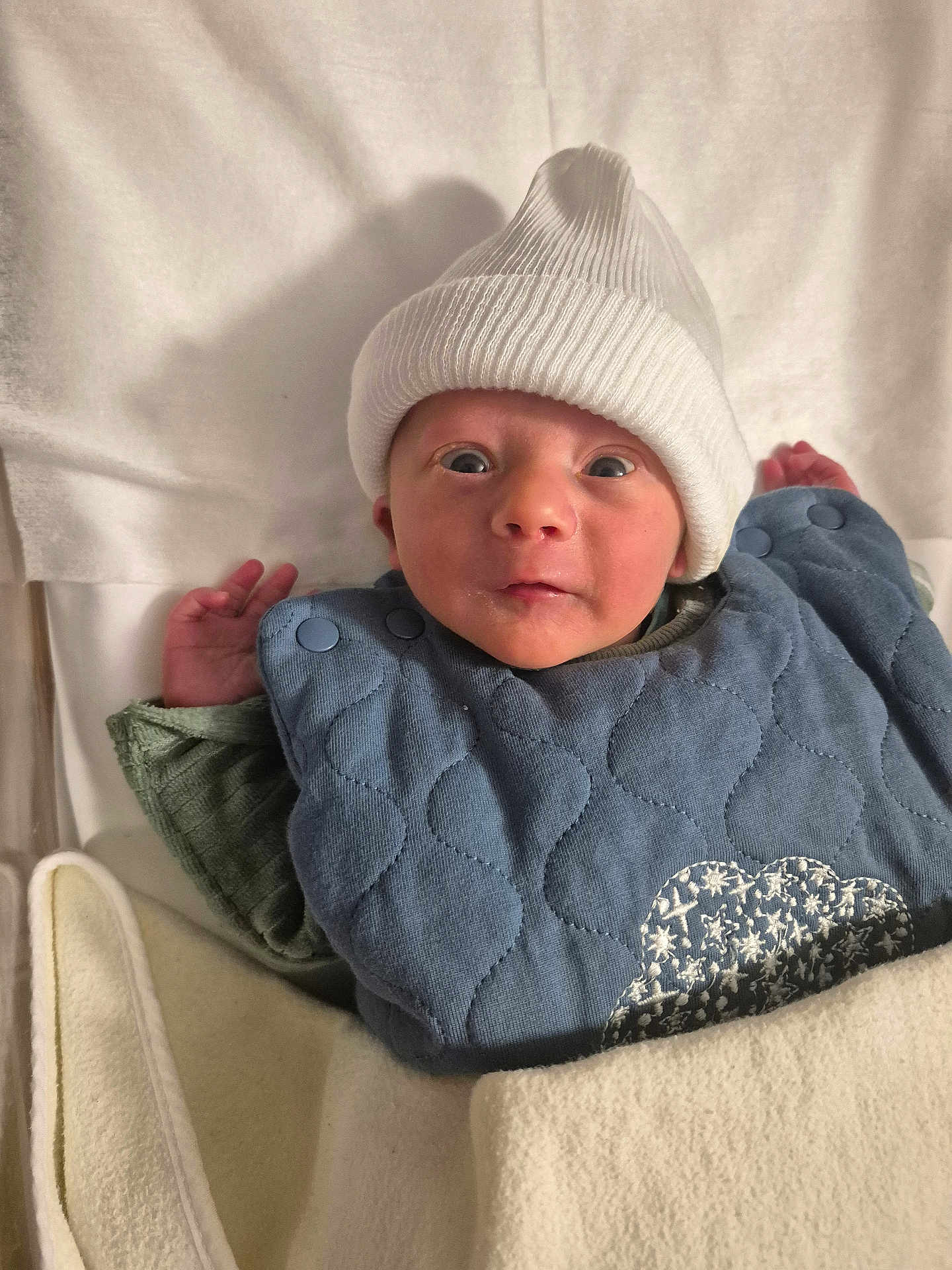 Livio participe au concours pour gagner de l'argent avec cette photo : baby, infant, hat, knit_hat, blue_bib, blanket, cozy, wide_eyes, curious, soft_texture, newborn, cute, child, lying_down, indoors, clothing, warm, hands, face, portrait