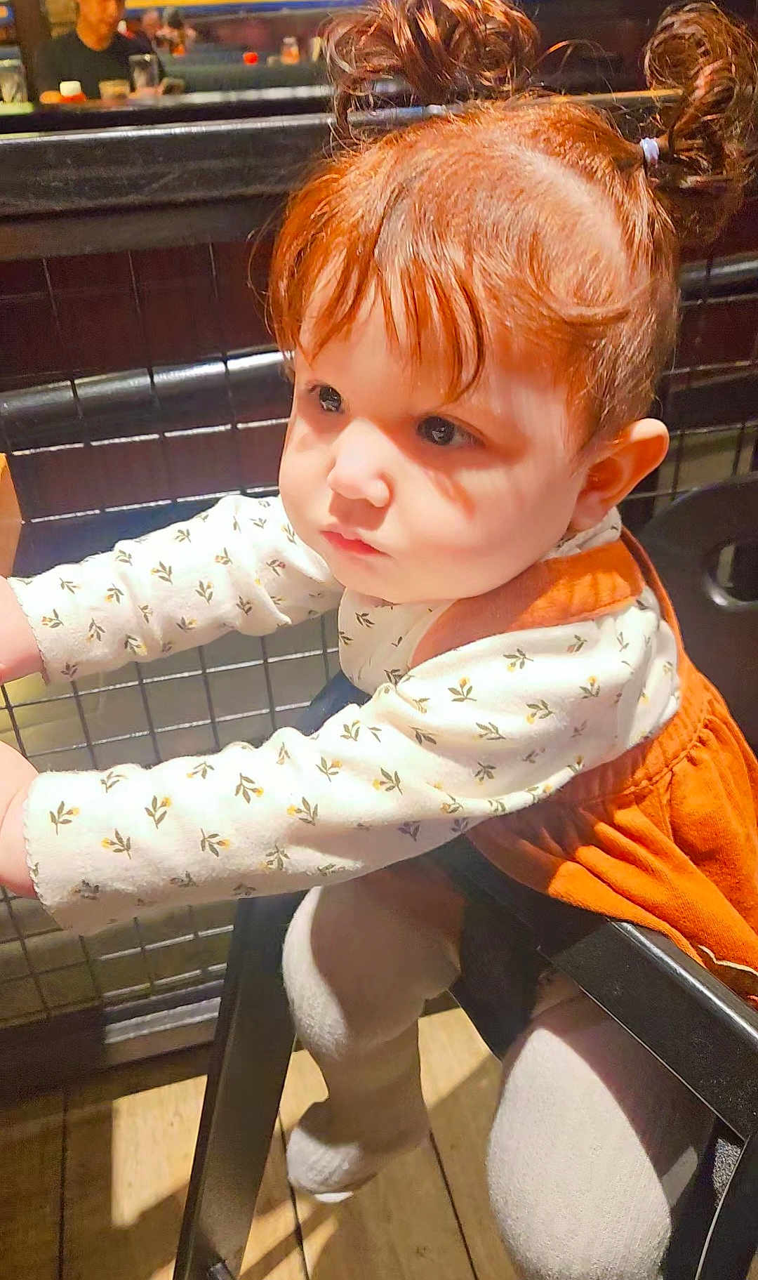 Alessandra joined the competition — help win amazing prizes! toddler, child, red_hair, pigtails, white_top, orange_dress, curious, indoor, restaurant, metal_grid, leaning, baby, person, cute, expression, seat, wooden_floor, clothing, face, portrait