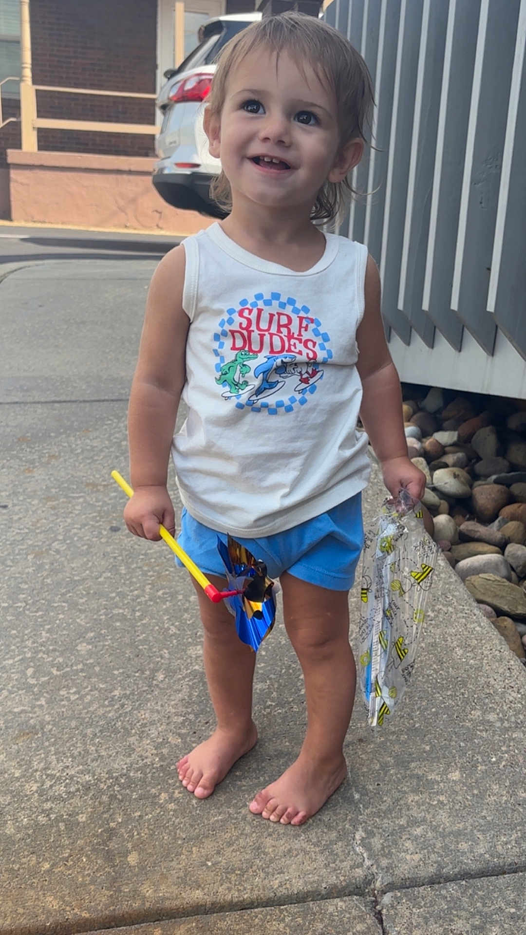 Anthony joined the competition — help win amazing prizes! toddler, child, barefoot, smiling, tank_top, shorts, pinwheel, plastic_bag, sidewalk, outdoor, happy, summer, playful, person, young_child, casual_clothing, daylight, curly_hair, cute, standing