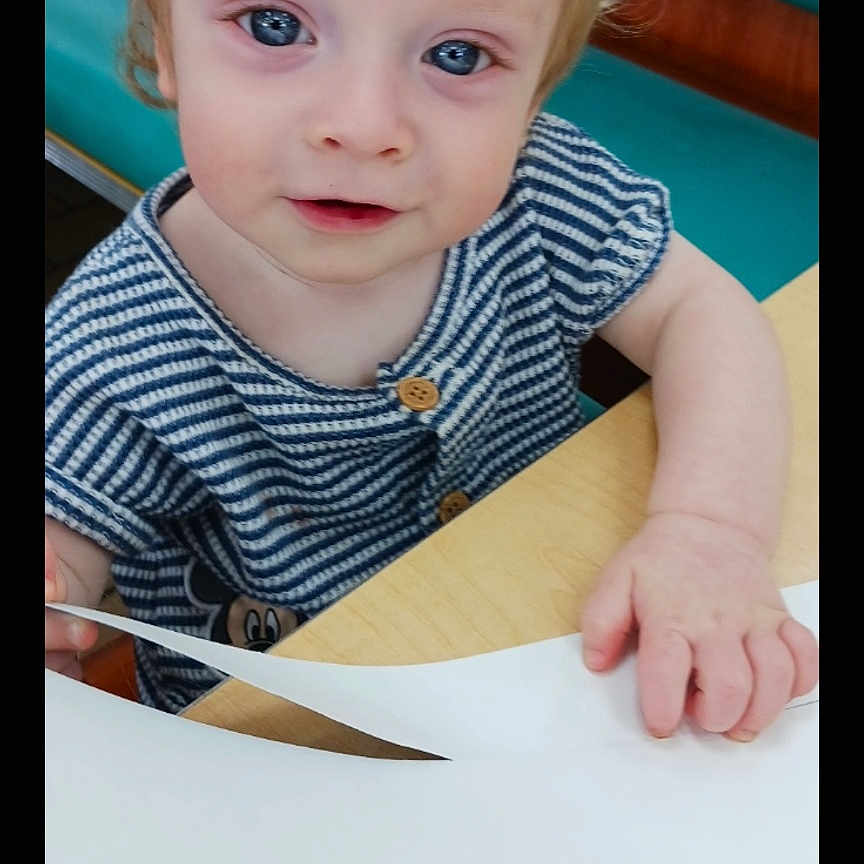 Camdyn is registered to the contest to win money with this photo: baby, bench, blonde_hair, blue_eyes, casual_clothing, child, curious, cute, face, hand, indoor, paper, playful, portrait, sitting, striped_shirt, table, toddler, wood, young_child