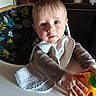 baby, child, high_chair, bib, toy, hand, face, indoor, kitchen, table, expression, clothing, seat, pattern, person, young_child, curious, light, colorful, furniture