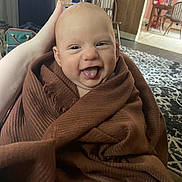 Weston is registered to the contest to win money with this photo: baby, blanket, smile, tongue, happy, wrapped, infant, hand, living_room, rug, furniture, wood_paneling, chair, home, cozy, person, indoor, cute, expression, skin
