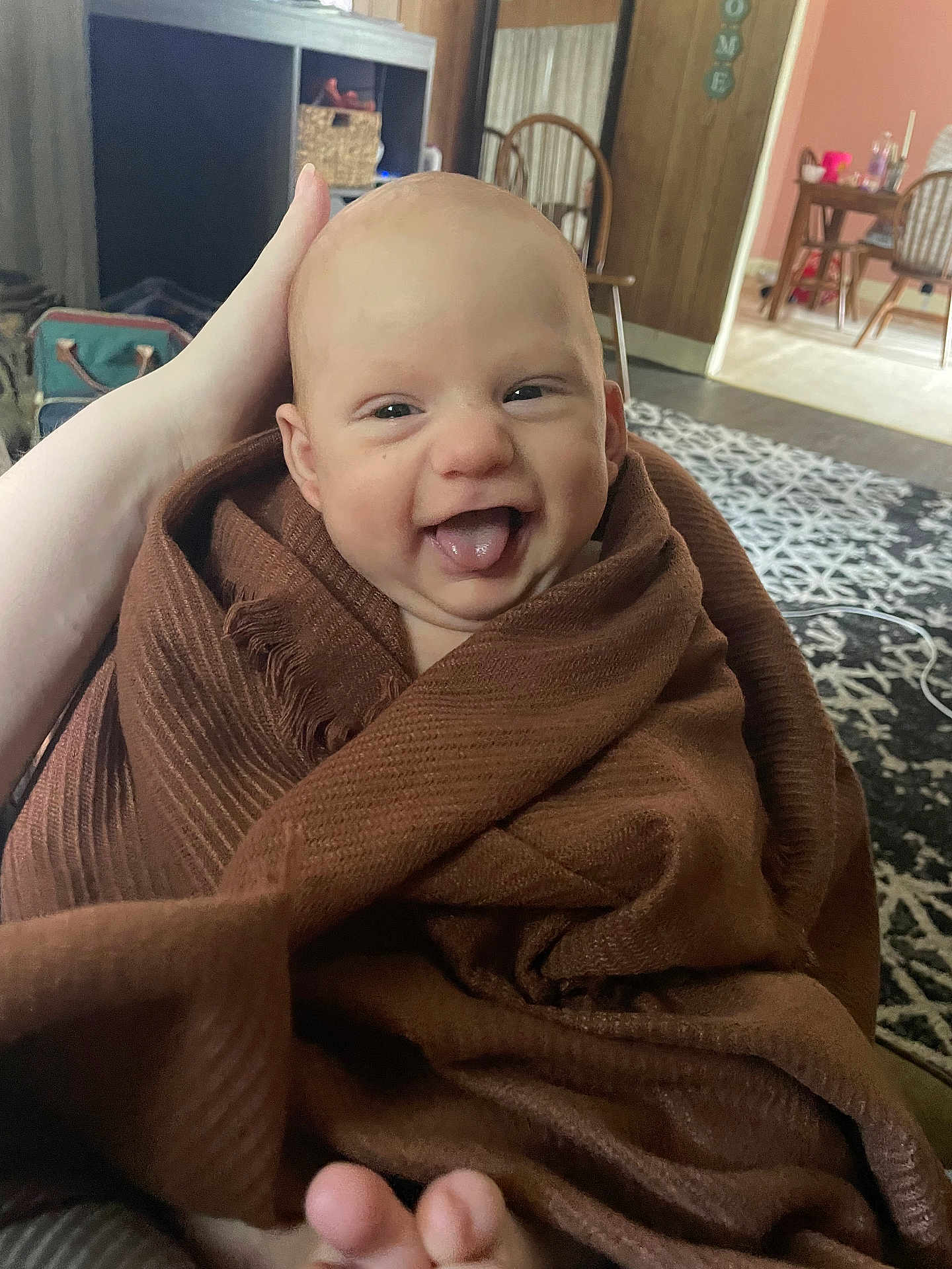 Weston is registered to the contest to win money with this photo: baby, blanket, smile, tongue, happy, wrapped, infant, hand, living_room, rug, furniture, wood_paneling, chair, home, cozy, person, indoor, cute, expression, skin