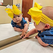 Alaina joined the competition — help win amazing prizes! baby, child, mirror, reflection, yellow_bow, headband, blue_clothing, table, indoor, curious, infant, skin, face, arms, hands, door, wall, person, young_child, expression