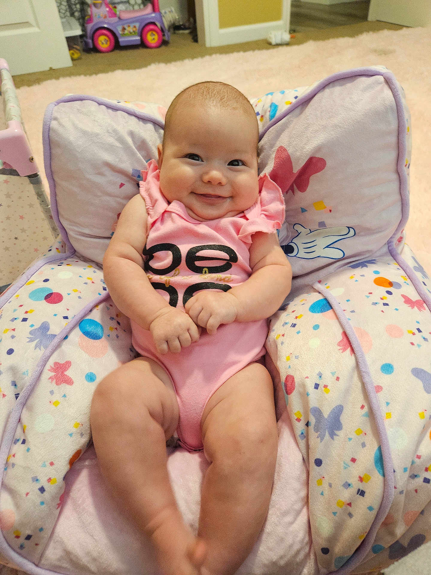 Alaina is registered to the contest to win money with this photo: baby, infant, child, pink_onesie, smiling, cushion, butterfly_pattern, indoor, toy_car, carpet, happy, chubby, seat, cute, person, skin, furniture, room, playroom, comfortable