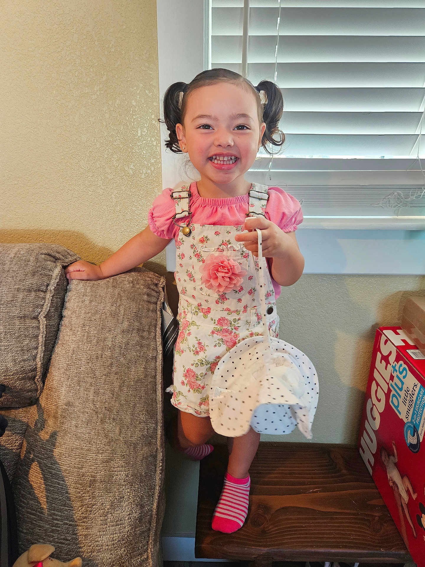 Juliette joined the competition — help win amazing prizes! child, girl, smiling, indoor, pigtails, pink_clothing, floral_overall, hat, socks, couch, window_blinds, wooden_bench, diaper_box, happy, person, portrait, home, furniture, cute, playful