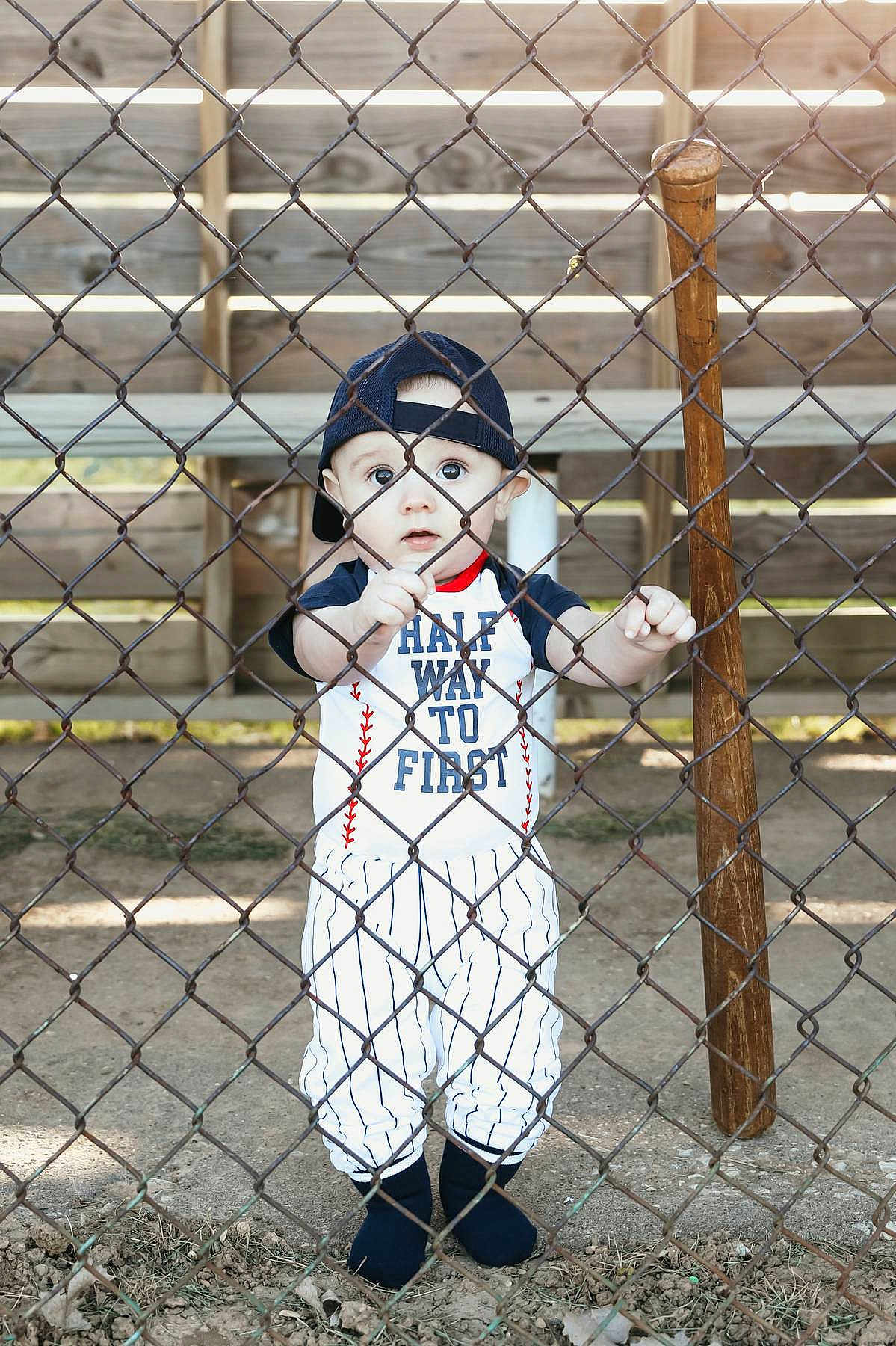 Beckham is registered to the contest to win money with this photo: baby, child, baseball, sports, fence, chain_link_fence, bat, bench, hat, cap, outdoor, dirt, clothing, person, young_child, cute, standing, portrait, daylight, curious