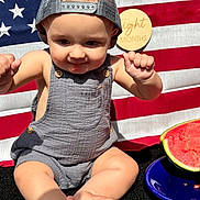 Beckham is registered to the contest to win money with this photo: baby, child, infant, cap, overalls, watermelon, plate, american_flag, milestone, sign, sitting, cute, summer, outdoor, food, fruit, skin, feet, hands, portrait