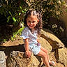 child, girl, smile, pigtails, rock, outdoor, sunlight, plants, greenery, flowers, nature, summer, happy, sitting, shorts, striped_shirt, sandals, daylight, portrait, young