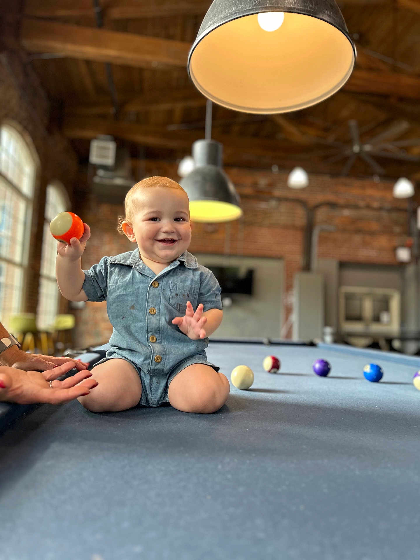 Leon is registered to the contest to win money with this photo: toddler, child, indoor, pool_table, ball, smiling, short_hair, denim_outfit, hands, lamp, brick_wall, window, game_room, furniture, happy, person, playing, kneeling, light, casual_clothing