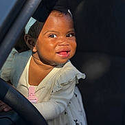 Londyn is registered to the contest to win money with this photo: toddler, child, smiling, face, headband, bow, dress, clothing, steering_wheel, car, vehicle_interior, happy, cute, young_child, portrait, human, baby, indoors, playful, curious