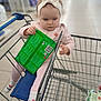 toddler, child, shopping_cart, grocery_store, headband, pink_clothing, socks, green_packet, floor, metal_cart, shopping, baby, seat, cute, indoor, person, retail, blurred_background, exploring, curious