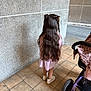 child, girl, long_hair, braids, pink_dress, white_shoes, standing, back_view, stroller, tiled_floor, stone_wall, outdoor, sidewalk, casual_clothing, young_child, hair_ties, shadow, urban, daylight, person
