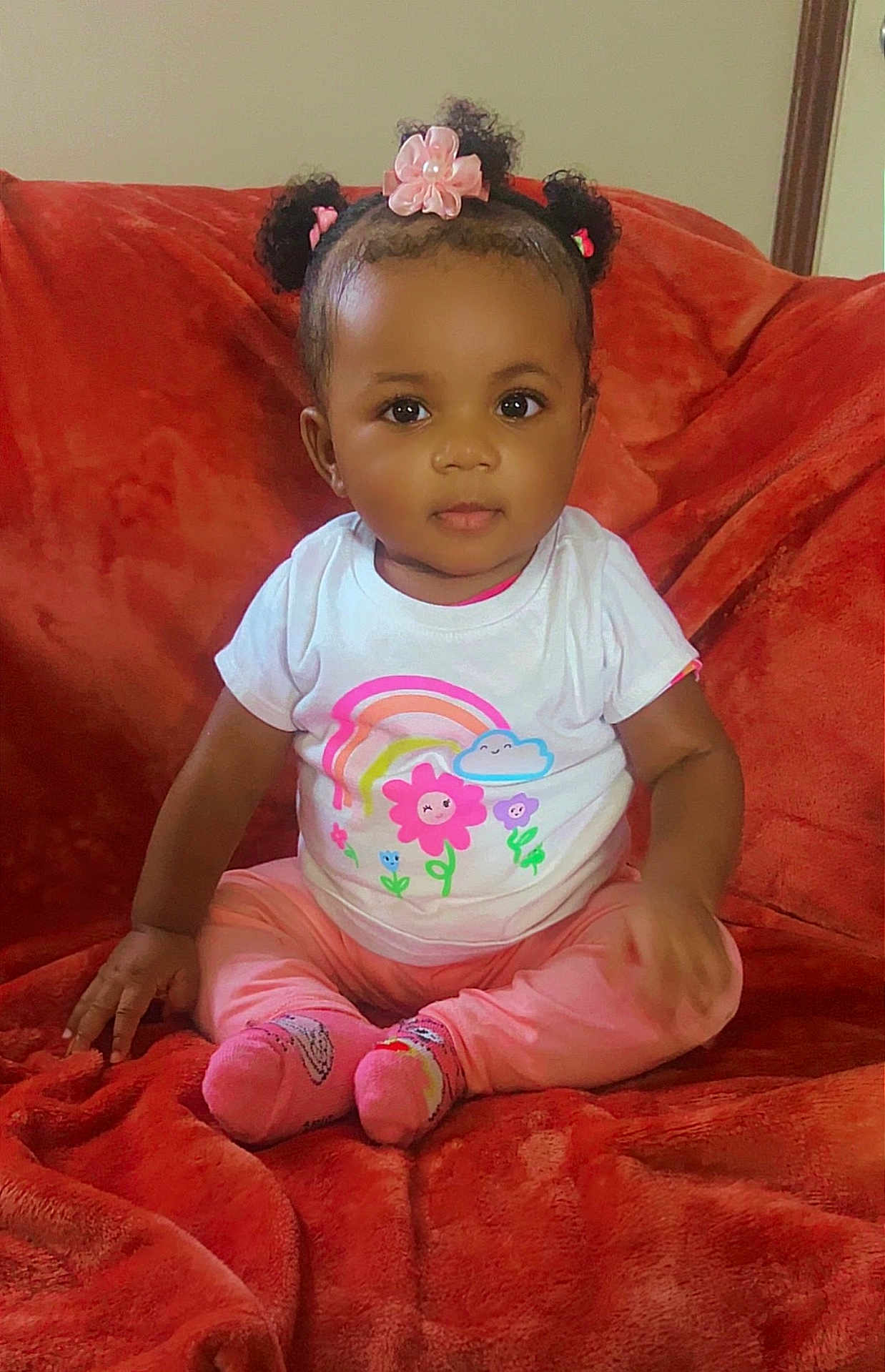 Zuri is registered to the contest to win money with this photo: baby, child, girl, pink, hair_accessories, sitting, white_shirt, rainbow, flowers, red_blanket, cute, indoors, portrait, smiling, happy, soft_texture, cozy, clothing, person, toddler