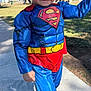 child, superman_costume, blue_suit, red_cape, yellow_belt, curly_hair, outdoor, sidewalk, grass, playground, smile, person, sunny, daytime, costume, superhero, cute, posing, footwear, park